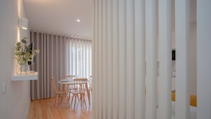 A modern interior with a wooden floor, a dining area with a round table and chairs, a white wall-mounted shelf with a vase, and a window with vertical blinds. - Liiiving in Porto Charming Downtown Delight I (Porto)