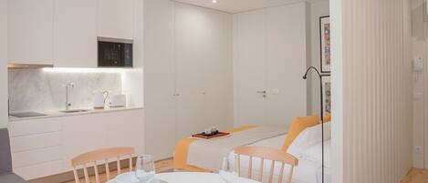 A modern, well-lit room with a round dining table set for two, a sofa with cushions, and a kitchen area with white cabinets and a microwave.