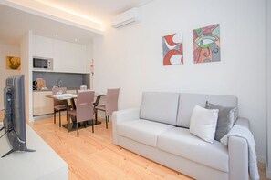 Ein modernes Wohnzimmer mit grauem Sofa, einem Esstisch mit Stühlen, einer Küchenzeile und zwei abstrakten Bildern an der Wand.