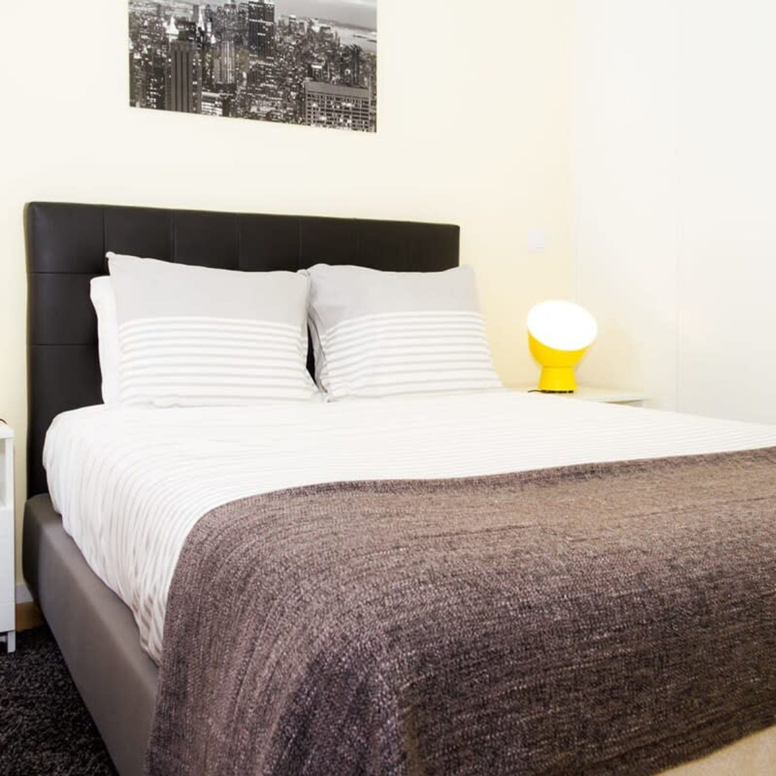 Cama bem arrumada com almofadas brancas e listradas, cabeceira escura e manta cinza.