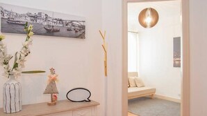 Et værelse med trægulv, en hvid kommode med skuffer, en vase med blomster, et billede på væggen, en høj gulvlampe og en døråbning til et andet værelse med en seng og en hængende lampe.