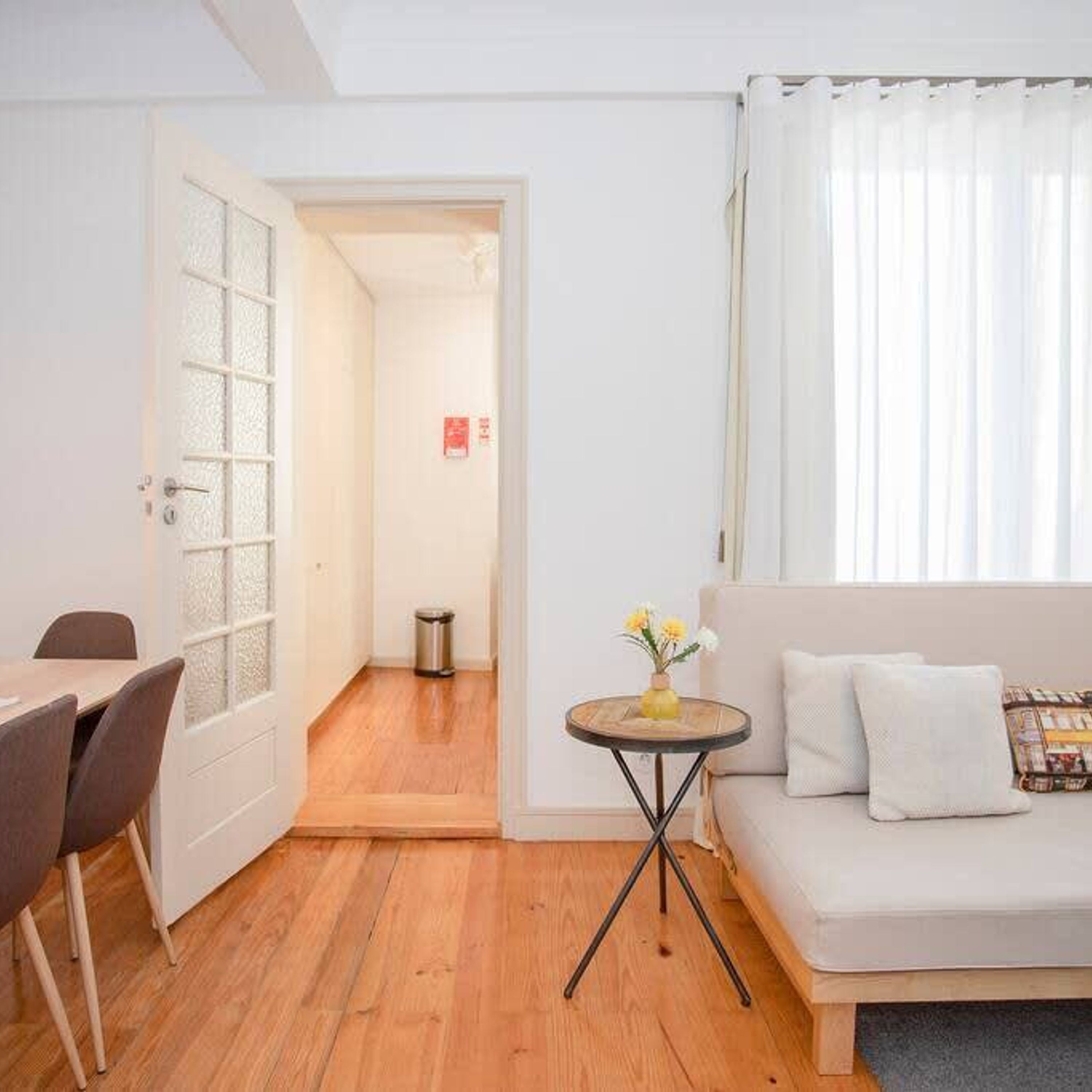 A room with a wooden floor, a dining table with chairs, a sofa, a small round table with a vase, and a door leading to another room.
