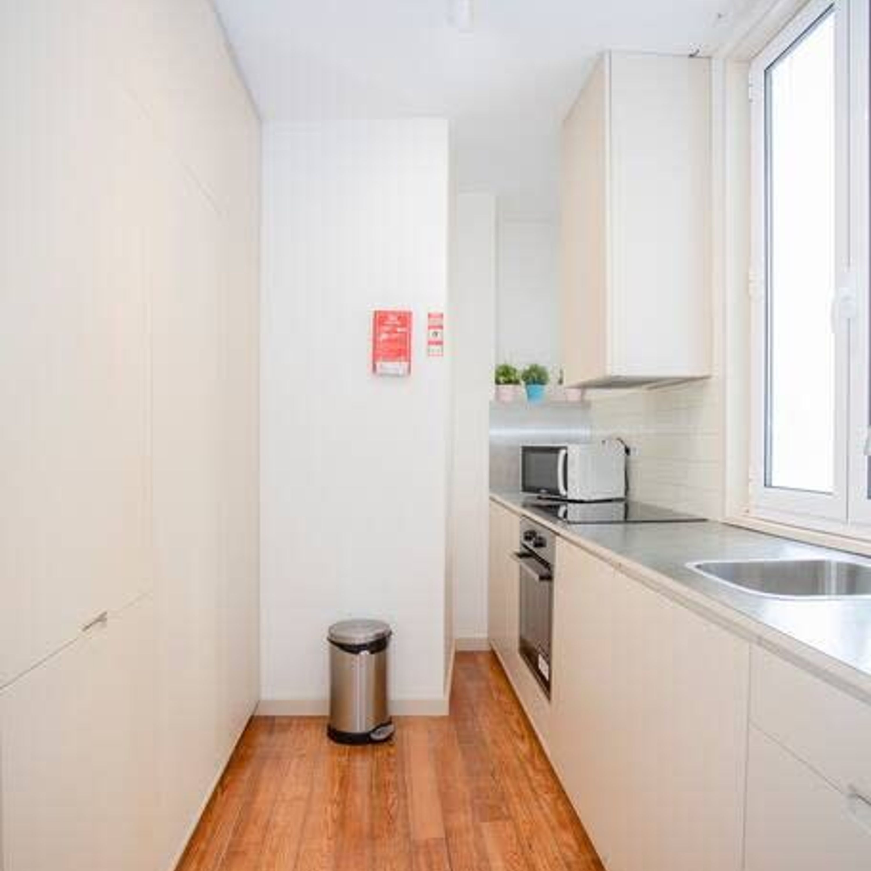 Een kleine keuken met witte kastjes, een roestvrijstalen spoelbak, een magnetron en een prullenbak.