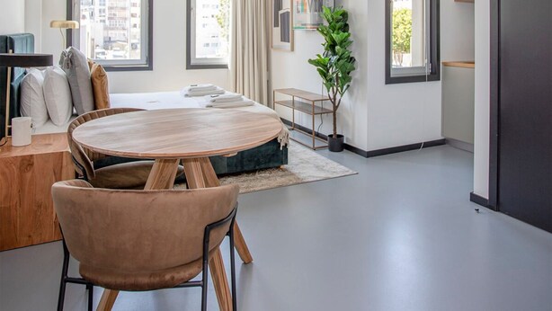 Egy modern, jól megvilágított nappali, melyben egy kerek, fából készült asztal, kényelmes ülőhelyek, nagy ablakok és minimalista berendezés található.