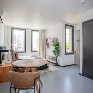 Een moderne, goed verlichte woonkamer met een ronde houten tafel, comfortabele zitplaatsen, grote ramen en een minimalistische inrichting.