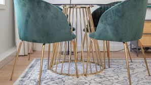 Une pièce avec une table ronde en verre, entourée de deux chaises de couleur turquoise aux pieds dorés, un lampadaire et une œuvre d’art abstraite encadrée accrochée au mur.