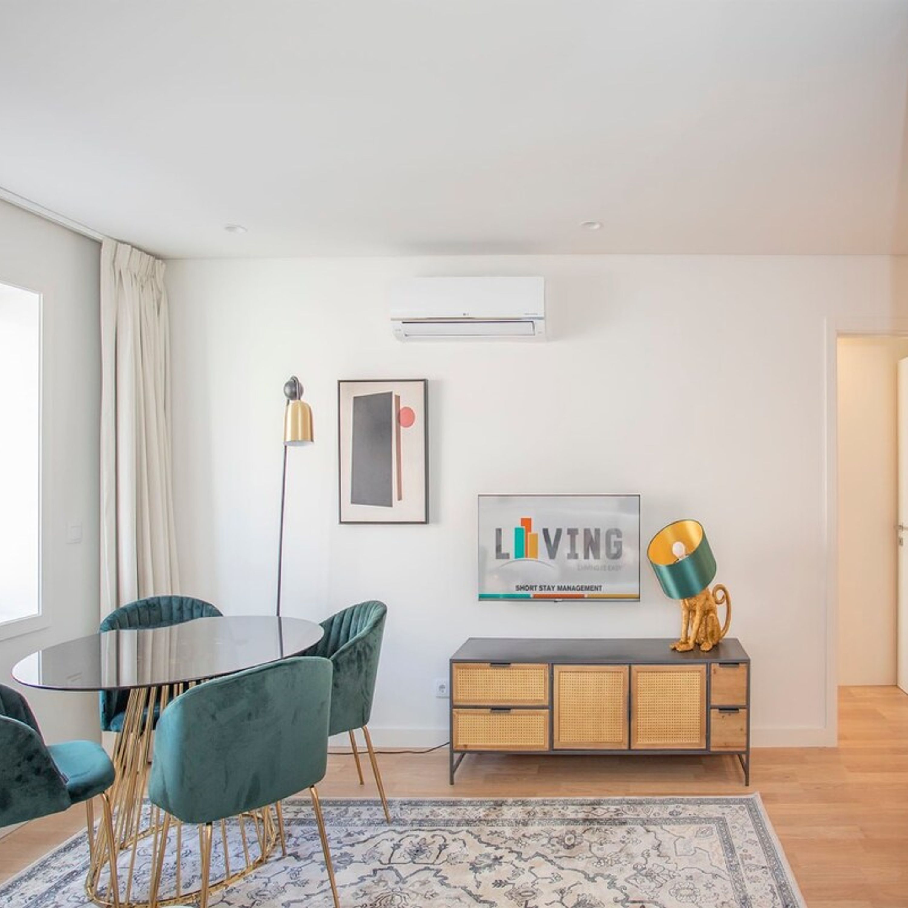 A modern living room with a round dining table, teal chairs, a rattan sideboard, and a mounted air conditioner.