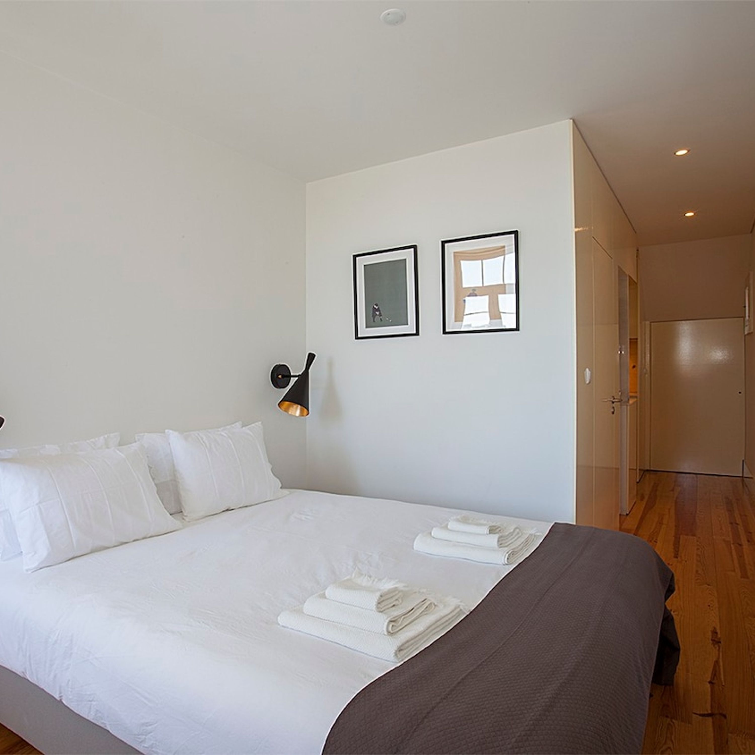 A hotel room with a large bed, white linens, a black wall-mounted lamp, and two framed pictures on the wall.
