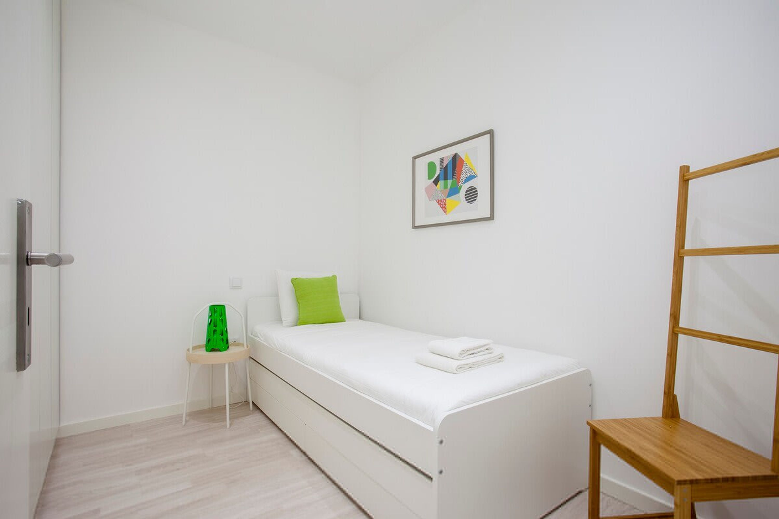 A small, white, single-bed room with a wooden chair, a small table, and a framed picture on the wall.