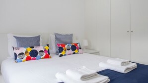 A neatly made bed with white and blue bedding, two colorful pillows, and a folded white towel on a blue bedspread. - Liiiving in Porto Santa Catarina Cosy Apartment (Porto)