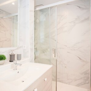 A modern bathroom with a marble wall, a glass shower enclosure, and a white sink with a plant and soap dispenser. - Liiiving in Matosinhos - Seaside Cosy Apartment (Matosinhos)