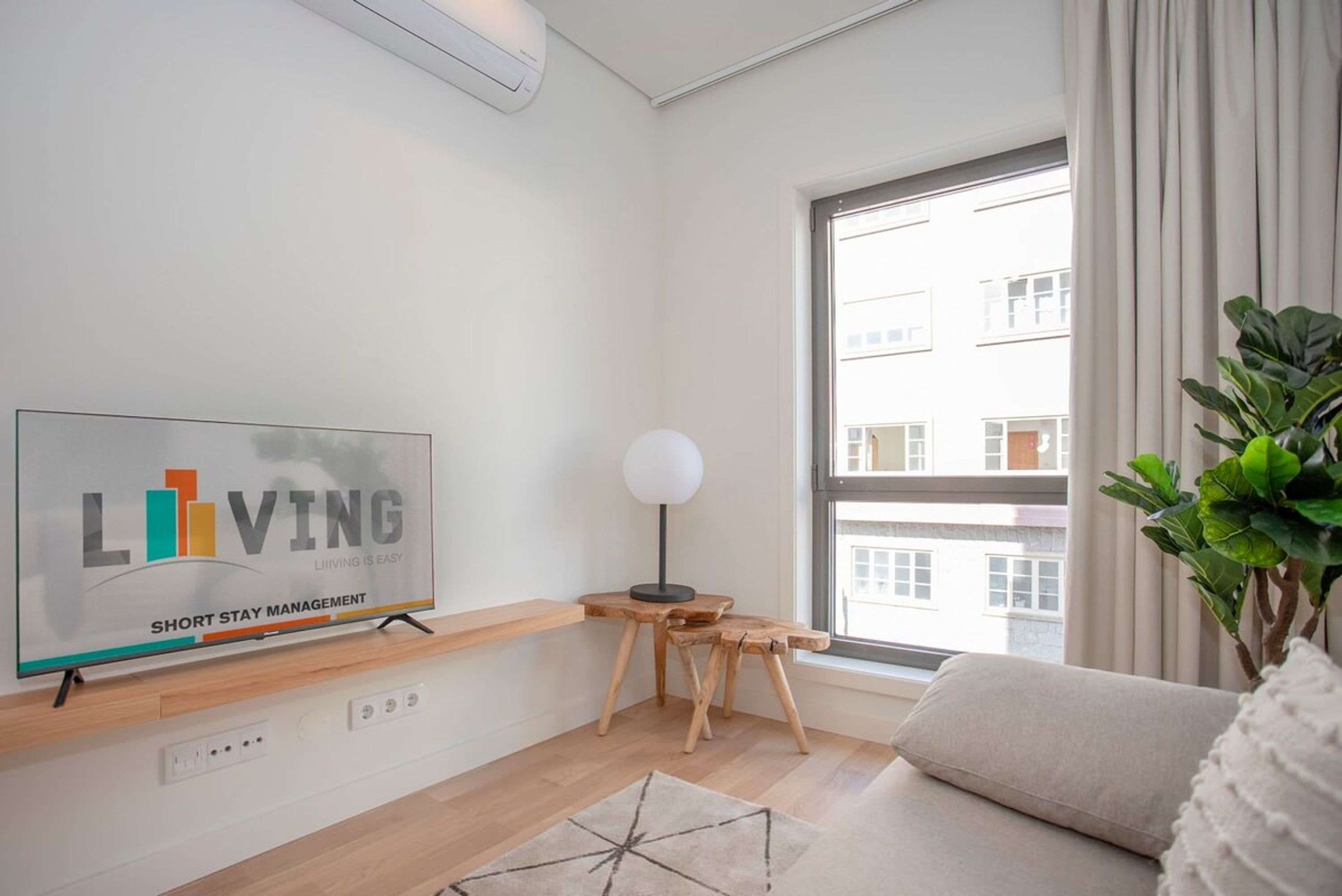 Un salon moderne comprenant une télévision, une petite table en bois, une lampe sur pied, un canapé et une grande fenêtre donnant sur des immeubles.