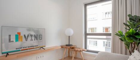 Un salon moderne comprenant une télévision, une petite table en bois, une lampe sur pied, un canapé et une grande fenêtre donnant sur des immeubles.