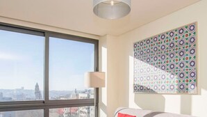 A modern living room with a large window, a grey sofa with red cushions, a glass coffee table, and a colorful patterned wall art. - Liiiving in Porto Downtown View Apartment (Porto)