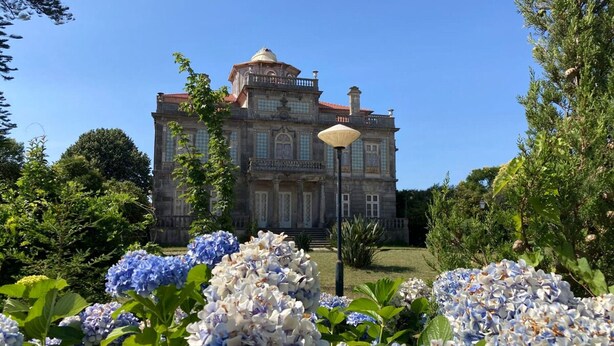Un grande edificio storico con una cupola centrale e numerose finestre, immerso nel verde e circondato da fiori di ortensia.