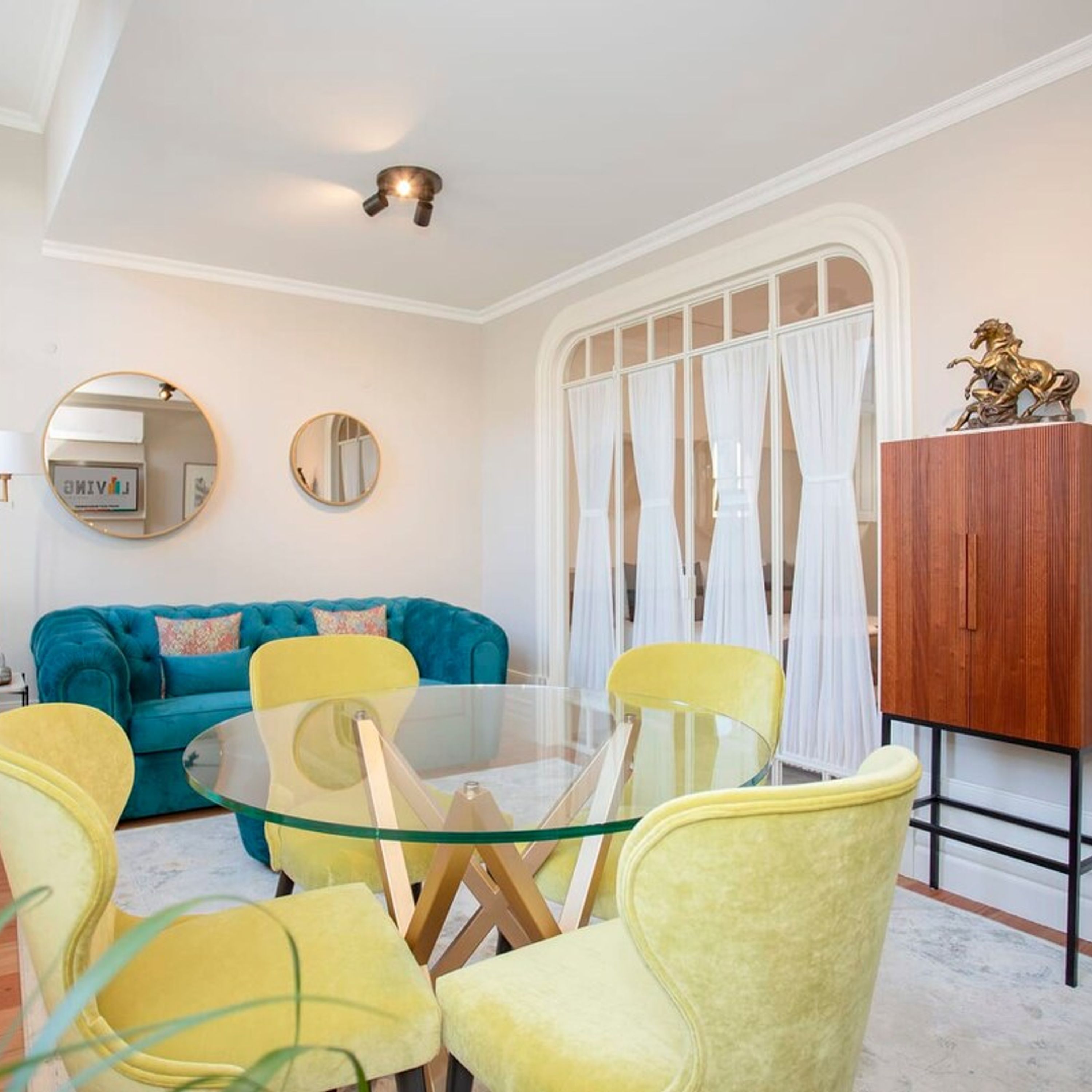 A modern living room with a glass table surrounded by yellow chairs, a blue sofa, and a wooden cabinet with a decorative horse statue.