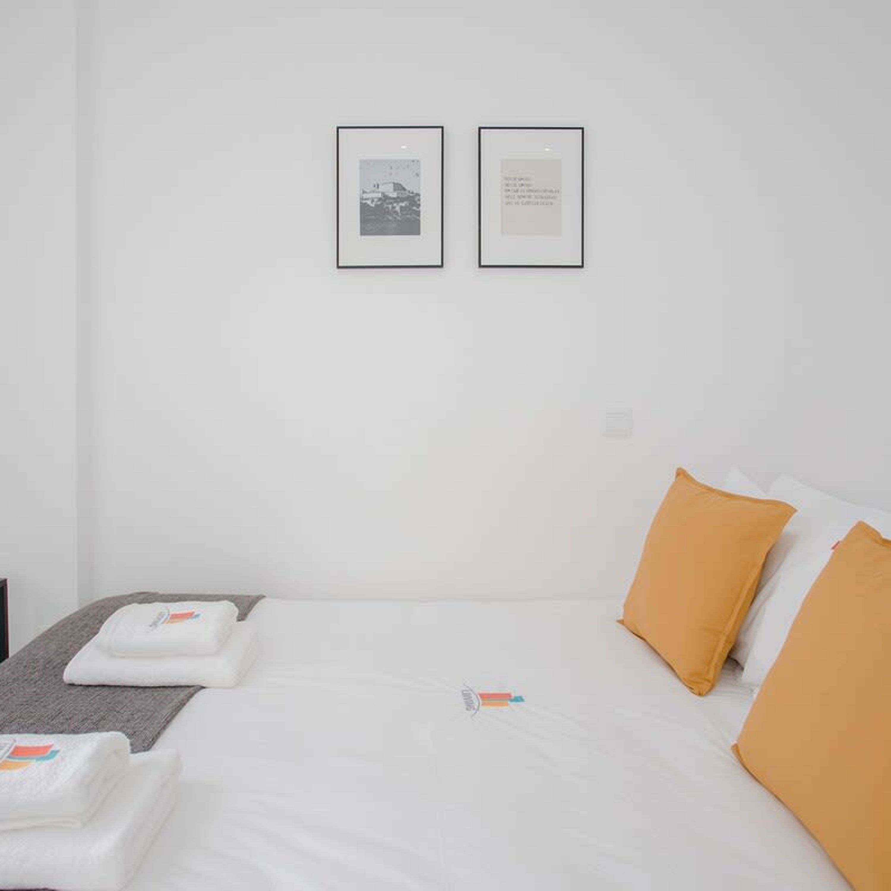 A minimalist hotel room with a neatly made bed, two framed pictures on the wall, and a small table with a towel and a bottle.