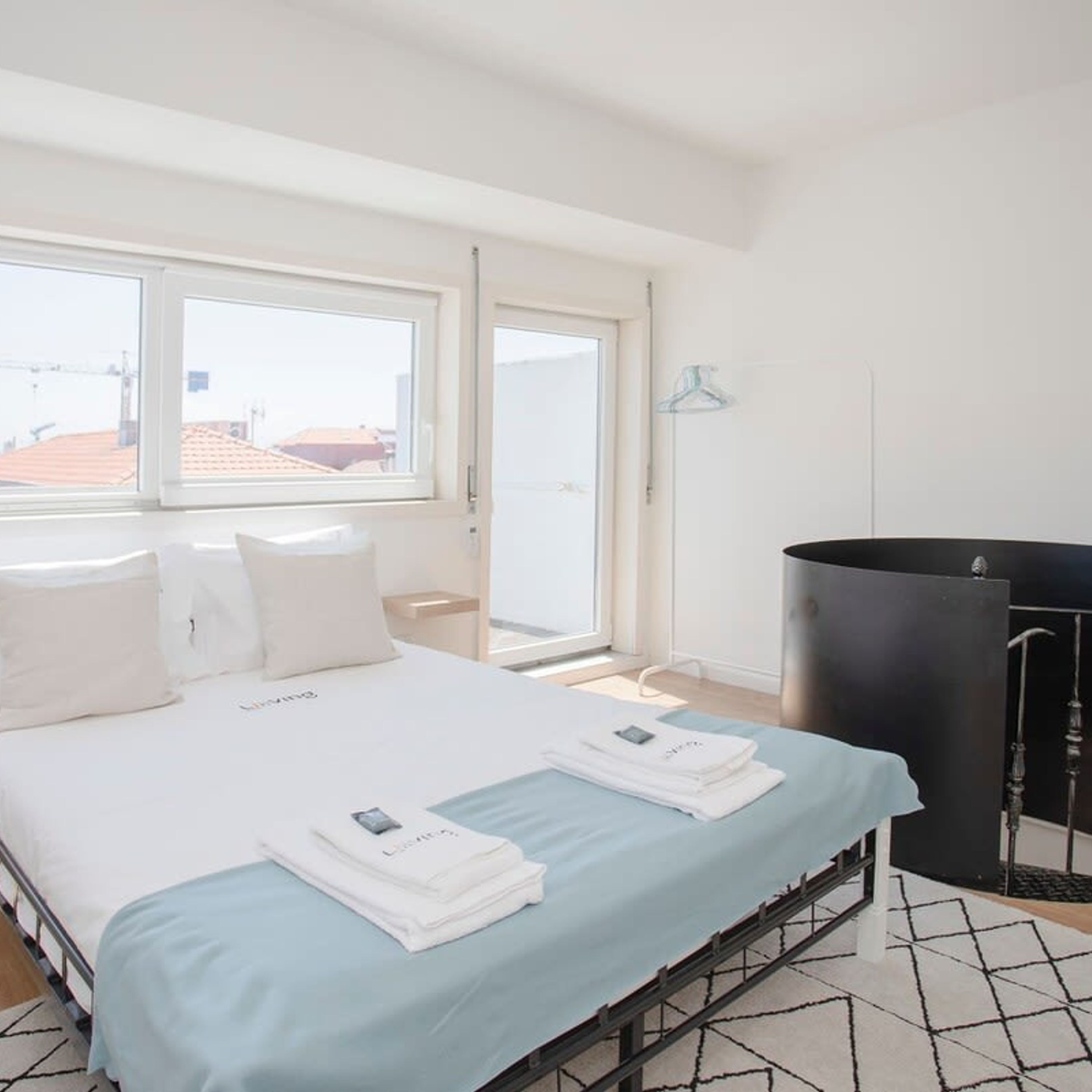 Une chambre d’hôtel avec un lit, une fenêtre donnant sur des immeubles, un radiateur noir et un mur blanc.