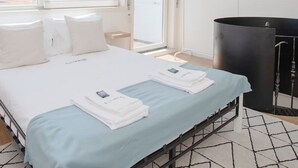 A hotel room with a bed, a window with a view of buildings, a black radiator, and a white wall. - Liiiving in Porto - Downtown Balcony Apartment (Porto)