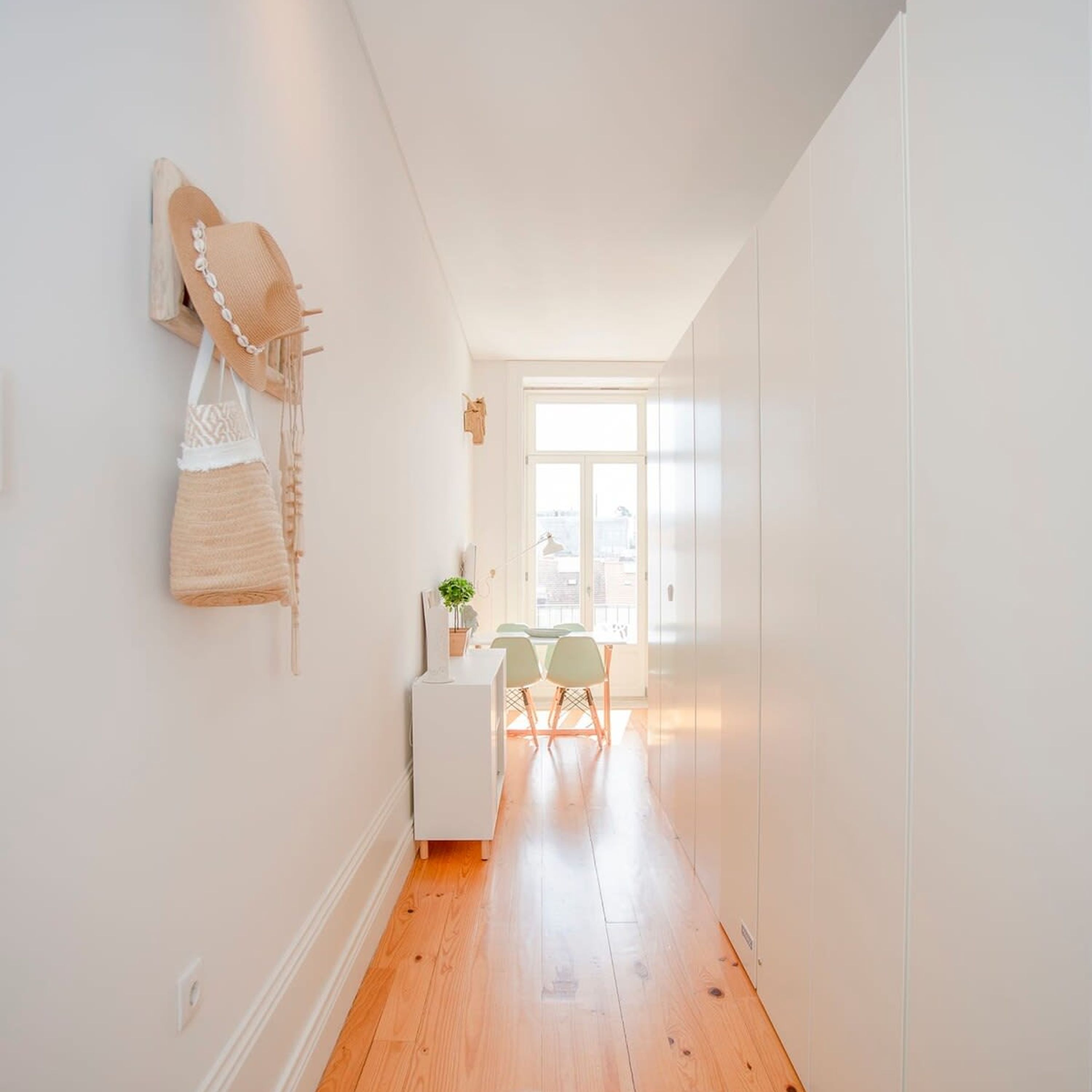 Un pasillo estrecho con suelo de madera, pared blanca y una ventana al final.