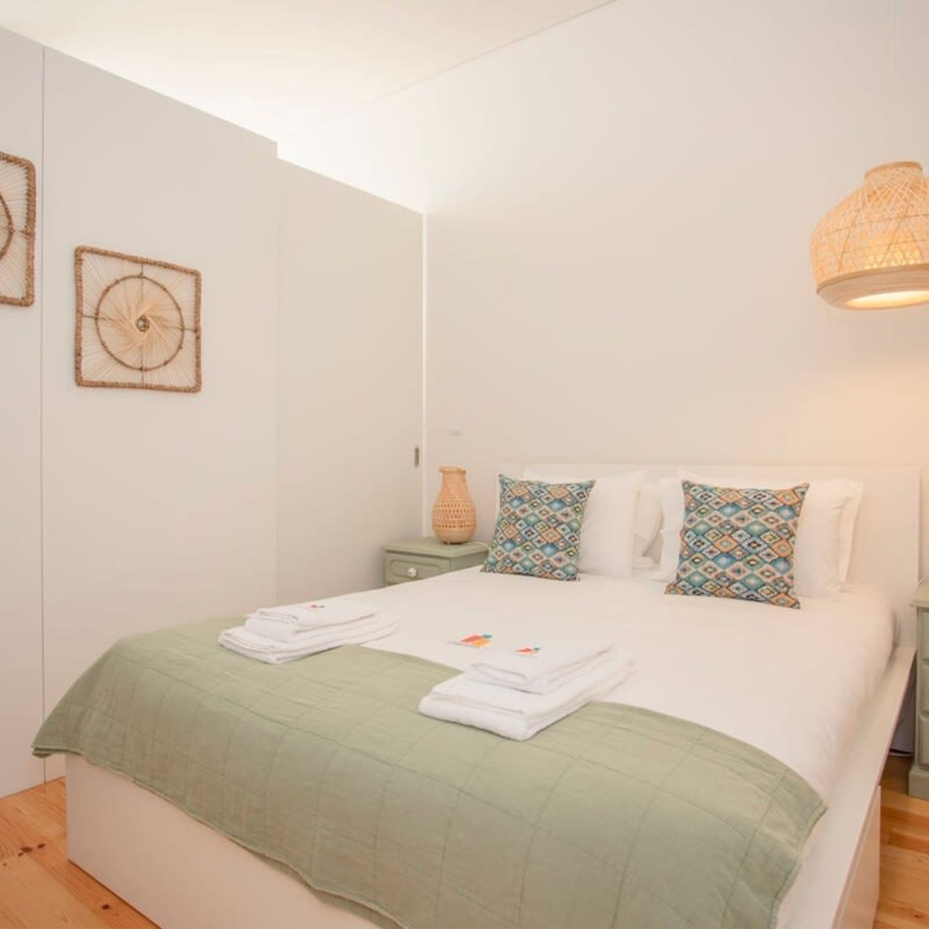 Un dormitorio con cama, ropa de cama verde, almohadas estampadas, una mesita de noche con lámpara y decoración en la pared.