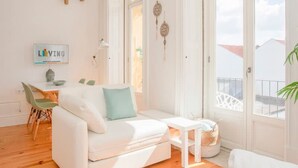 A bright living room with a white sofa, wooden flooring, a small table, and a view of the outside. - Liiiving in Porto - Downtown Balcony Studio (Porto)