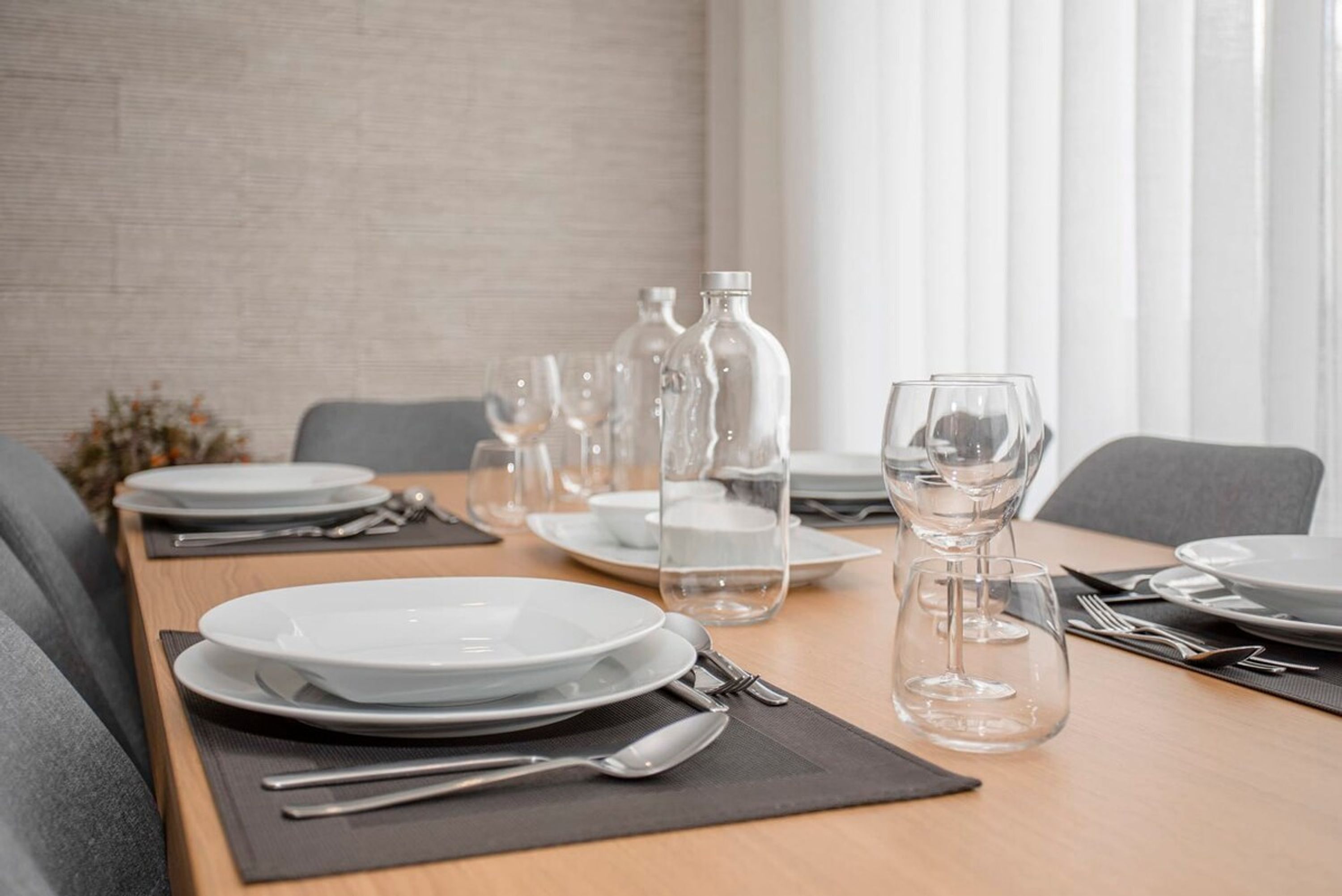 Une table à manger dressée avec des assiettes, des couverts et des verres, avec des bouteilles et un vase en arrière-plan.