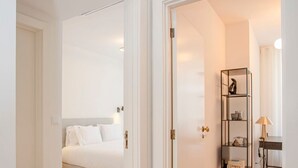 Ein Flur mit Holzboden, einem Bett mit weißer Bettwäsche und einem schwarzen Metallregal mit Dekoration.