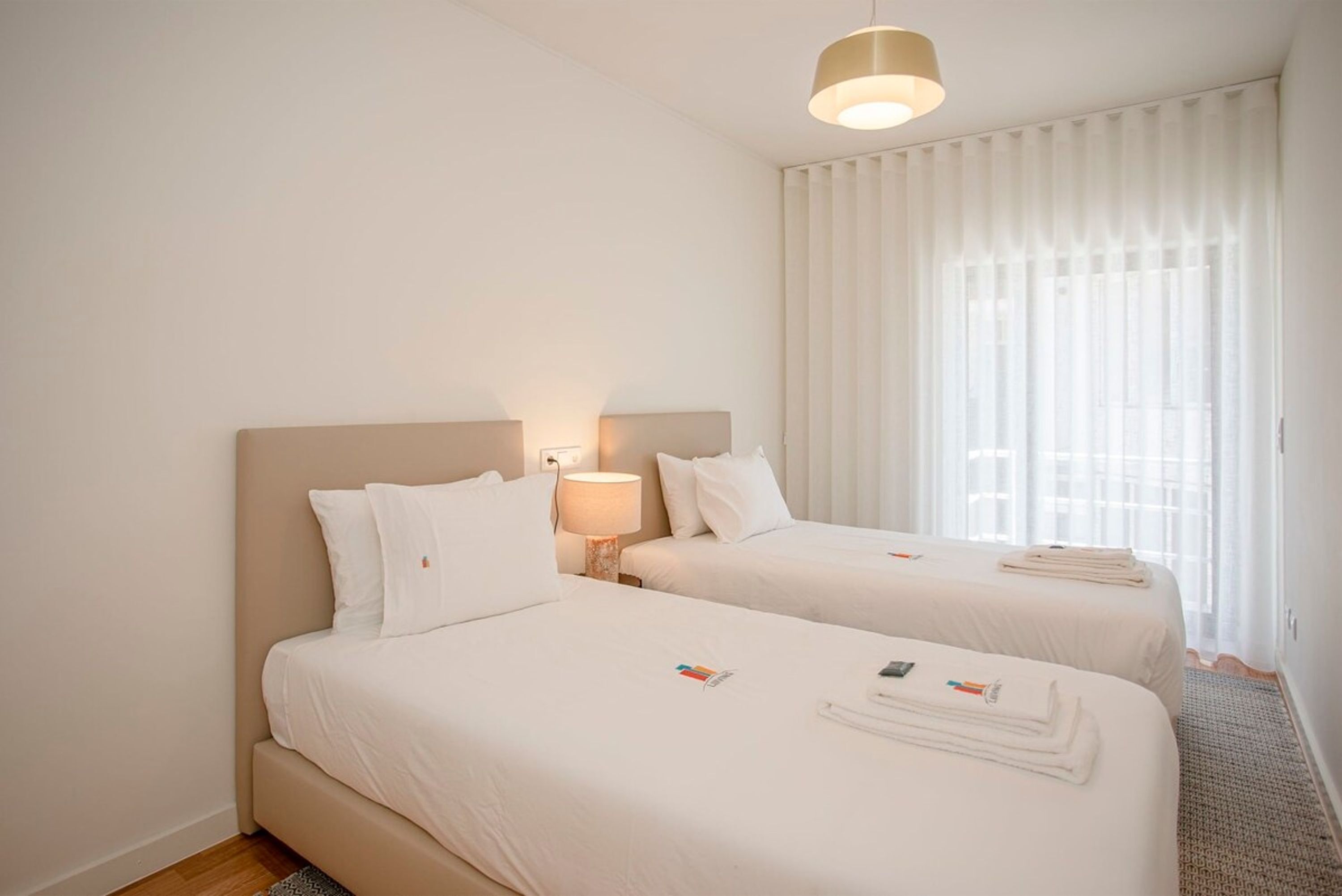 Une chambre d’hôtel avec deux lits, une lampe de chevet et une fenêtre avec des rideaux légers.