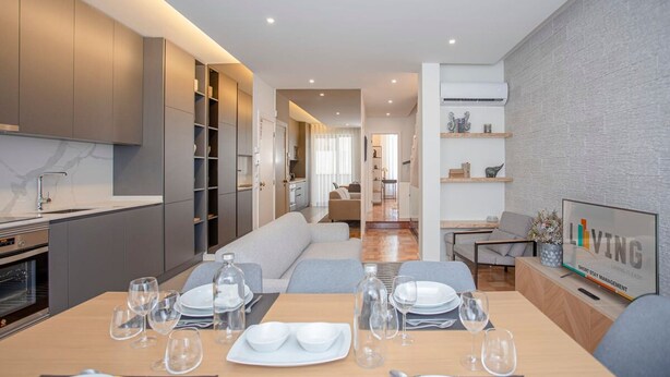 Eine moderne Küche mit integrierten Geräten, eine Essecke mit einem Holztisch, gedeckt für vier Personen, und ein Wohnbereich mit Sofa und Fernseher.