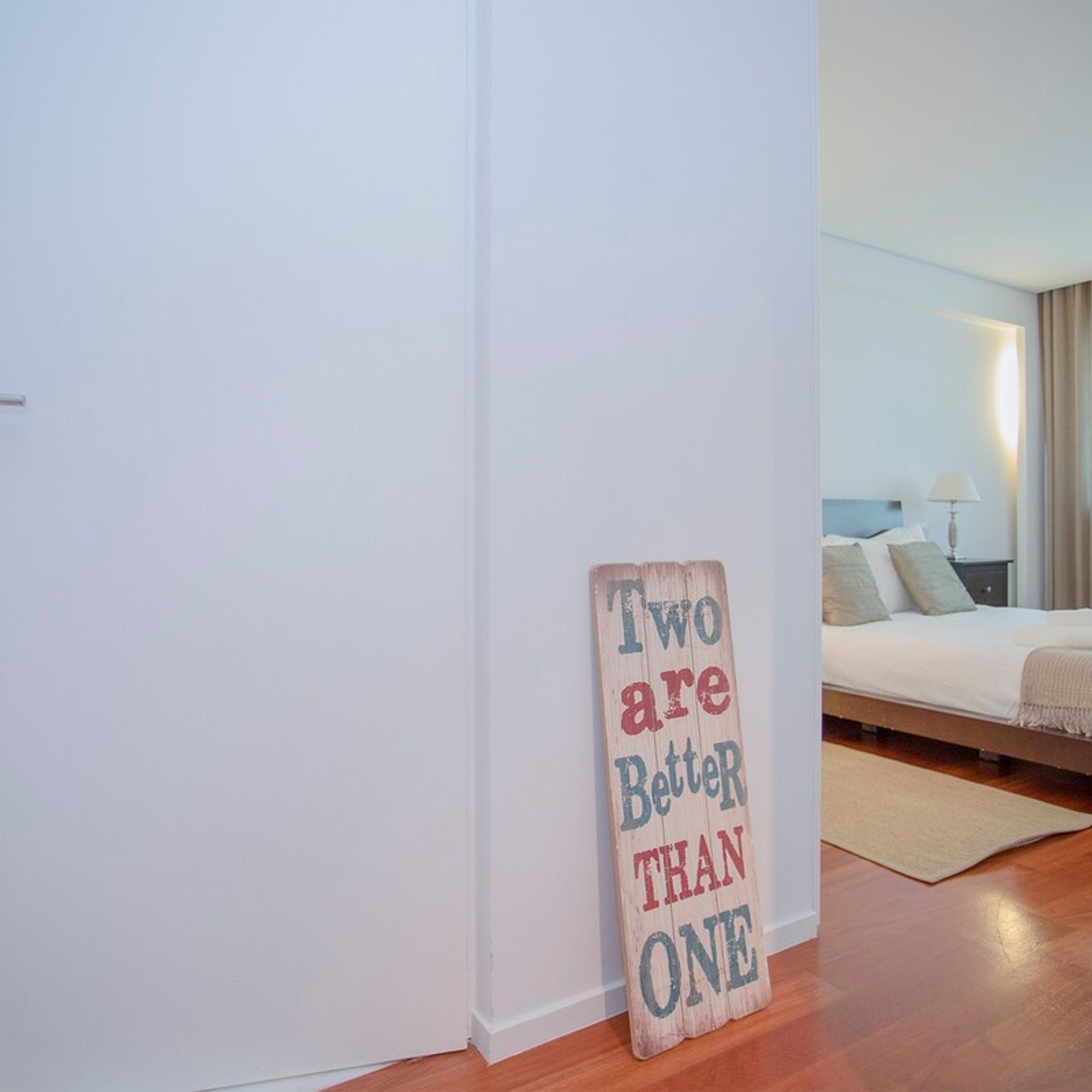 A bedroom with a bed, a bedside table, and a wall-mounted sign that reads "Two are Better Than One".