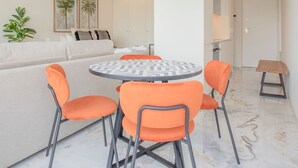 Un salon moderne avec une table ronde, des chaises orange et un canapé. Deux tableaux encadrés ornent le mur, ainsi qu’une plante et un luminaire suspendu.