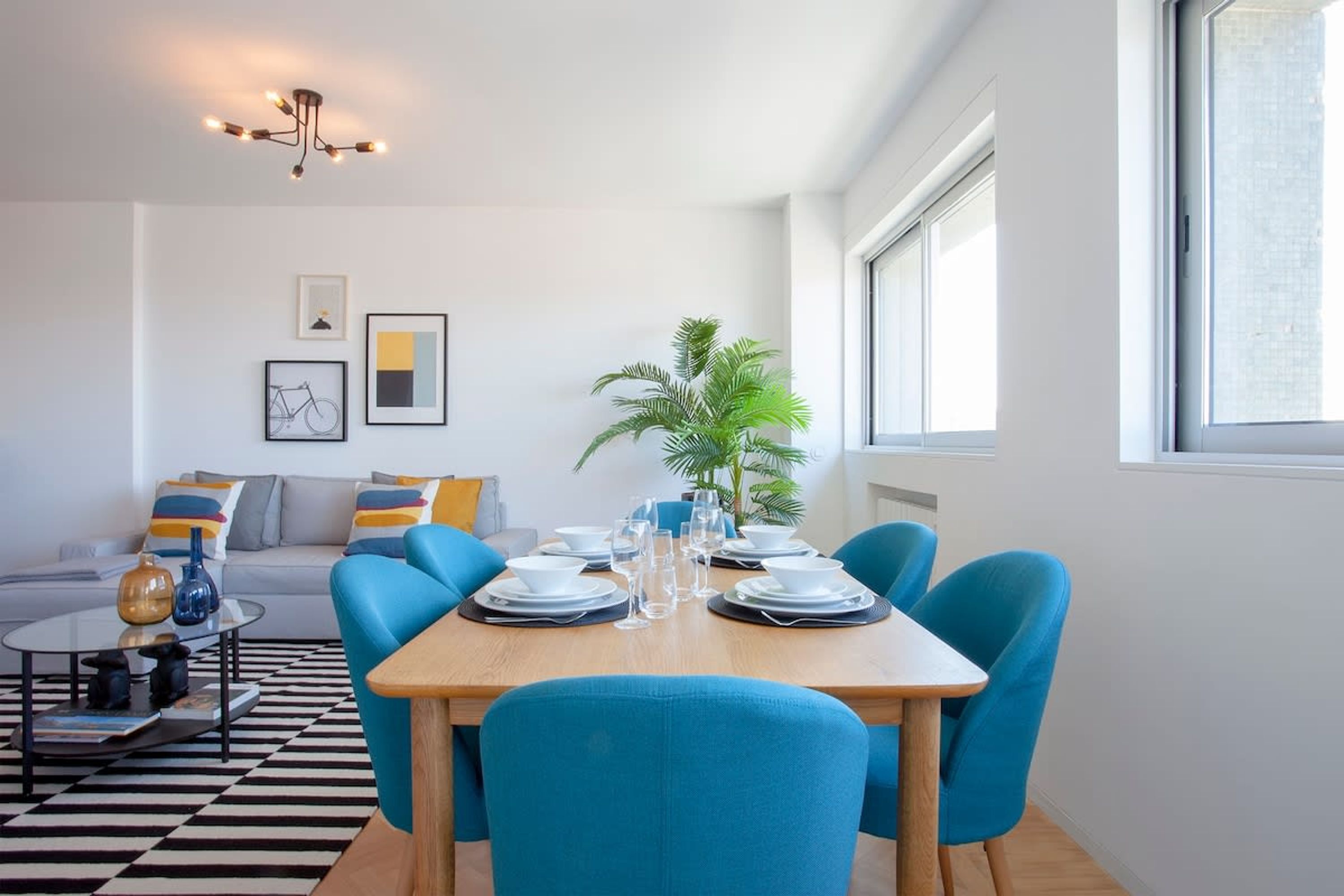 A modern dining area with a wooden table set for four, surrounded by blue chairs. There is a sofa with cushions in the background, a small round table with a black and white checkered table runner, and a potted plant to the side.