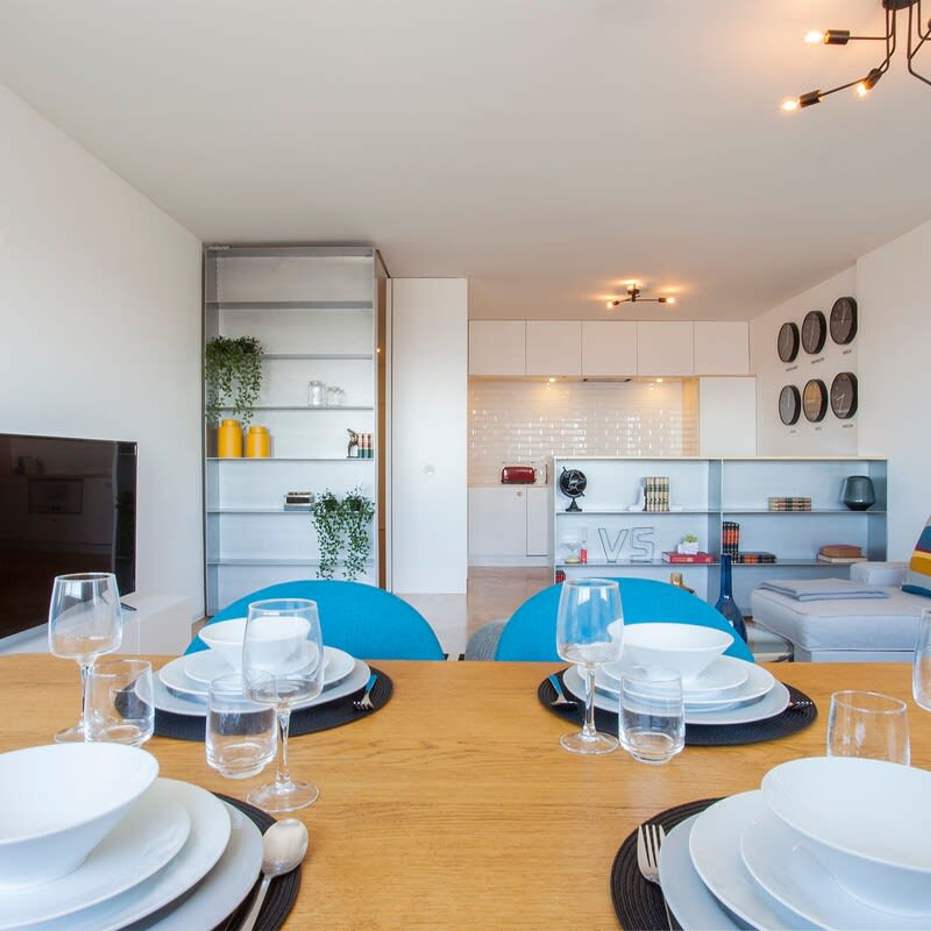 Ein moderner Essbereich mit Holztisch, gedeckt für eine Mahlzeit, eine Küche im Hintergrund und eine Wohnarea mit Sofa und Fernseher.