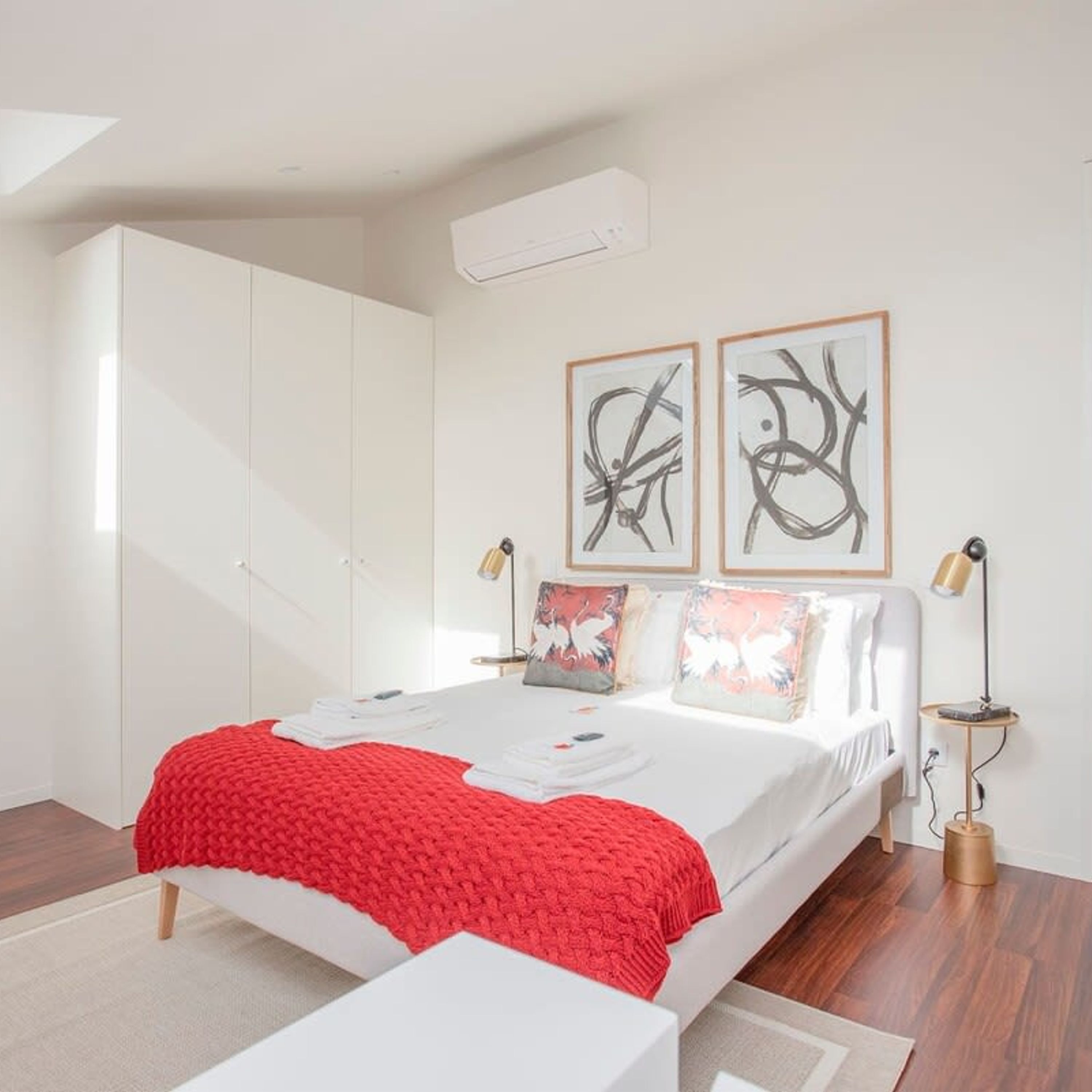 Ein modernes Schlafzimmer mit Bett, weißem Kleiderschrank, abstrakter Wandkunst und roter Decke.