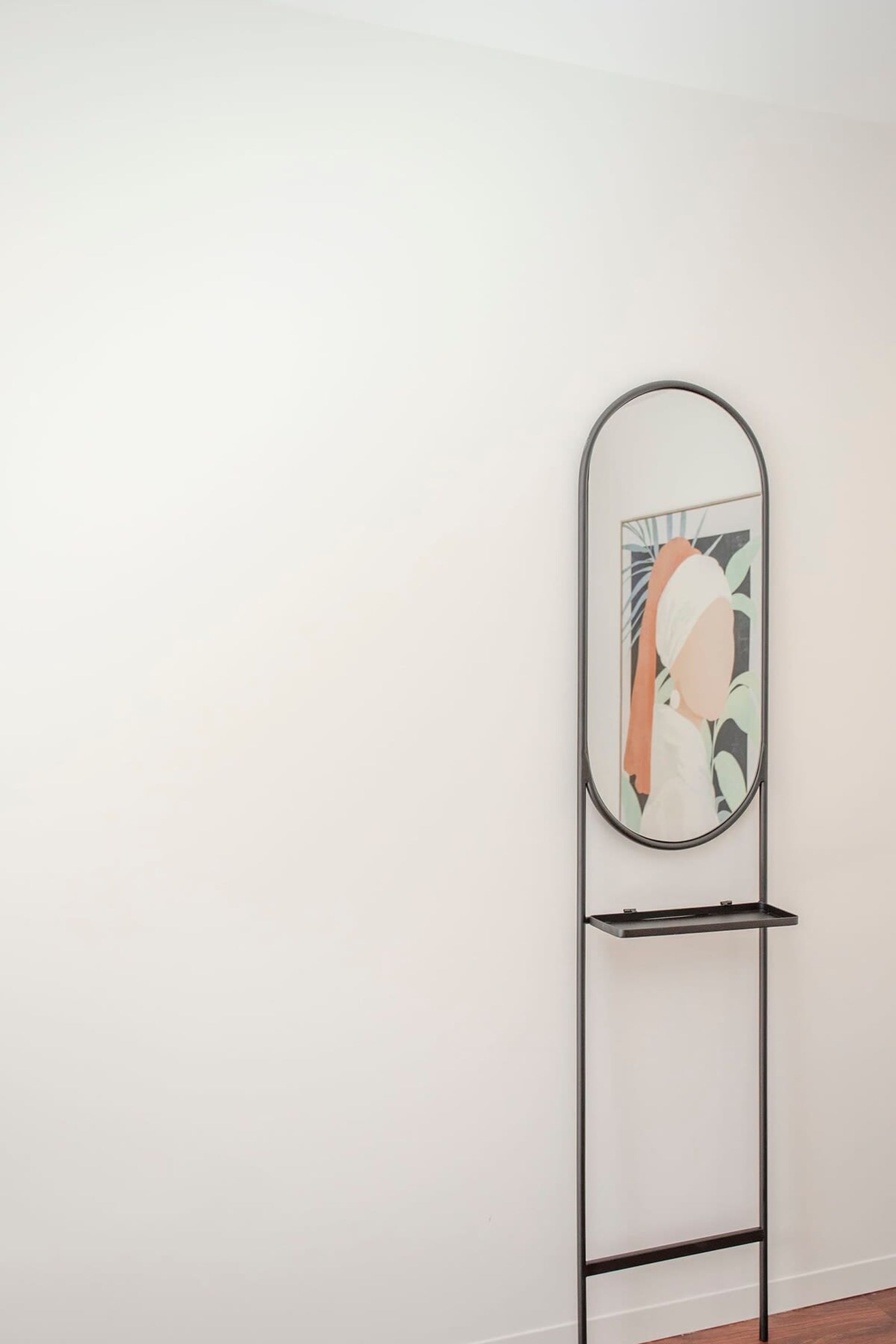 Un grand miroir autoportant au design minimaliste, fixé sur un socle en métal noir devant un mur blanc uni.