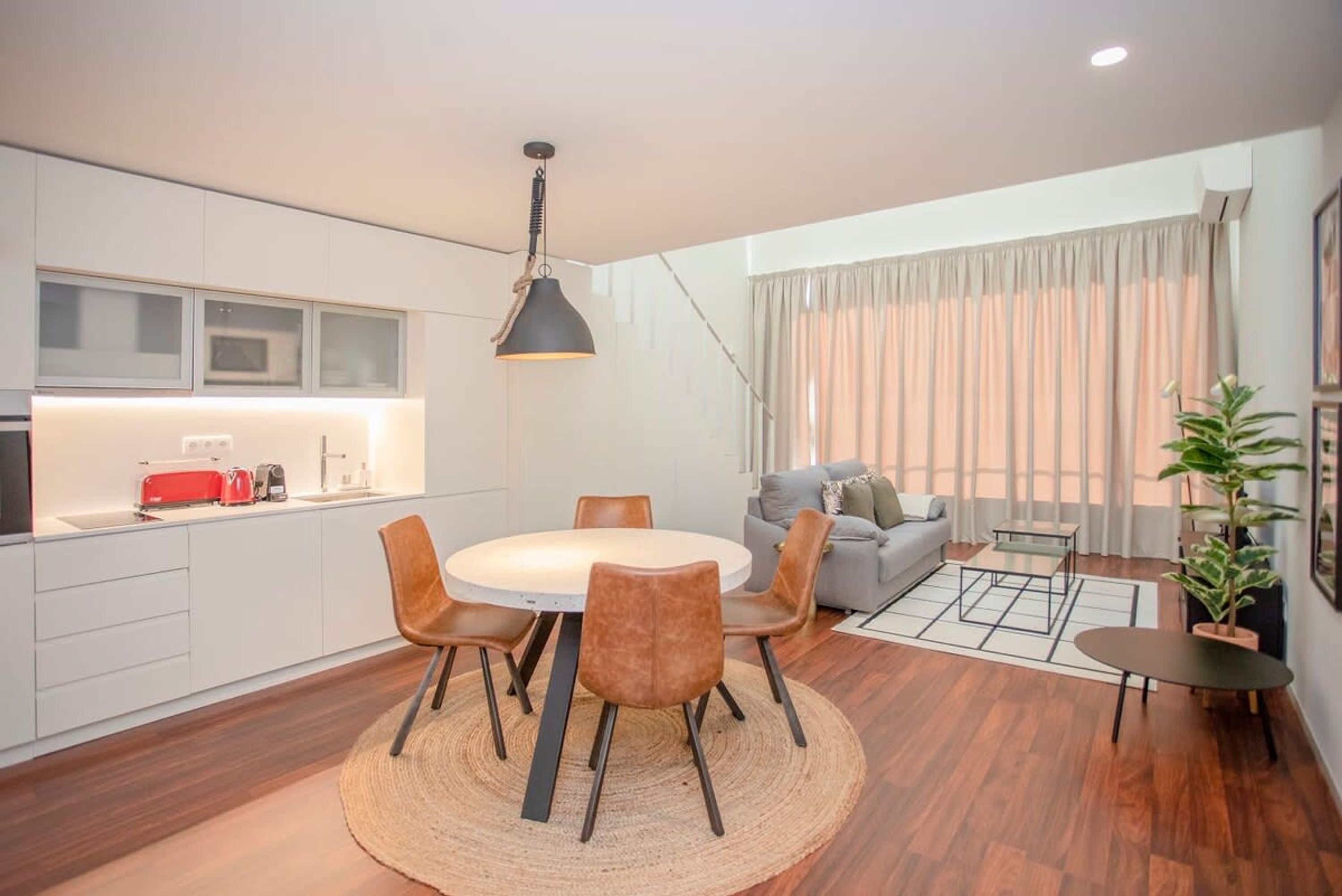 A modern kitchen with white cabinets, a round dining table with chairs, a hanging pendant light, and a grey sofa in the living area.