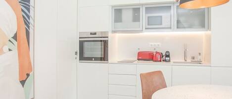 Una cocina moderna con armarios blancos, un microondas y una tostadora roja. Hay una mesa de comedor redonda con sillas marrones y una lámpara colgante.
