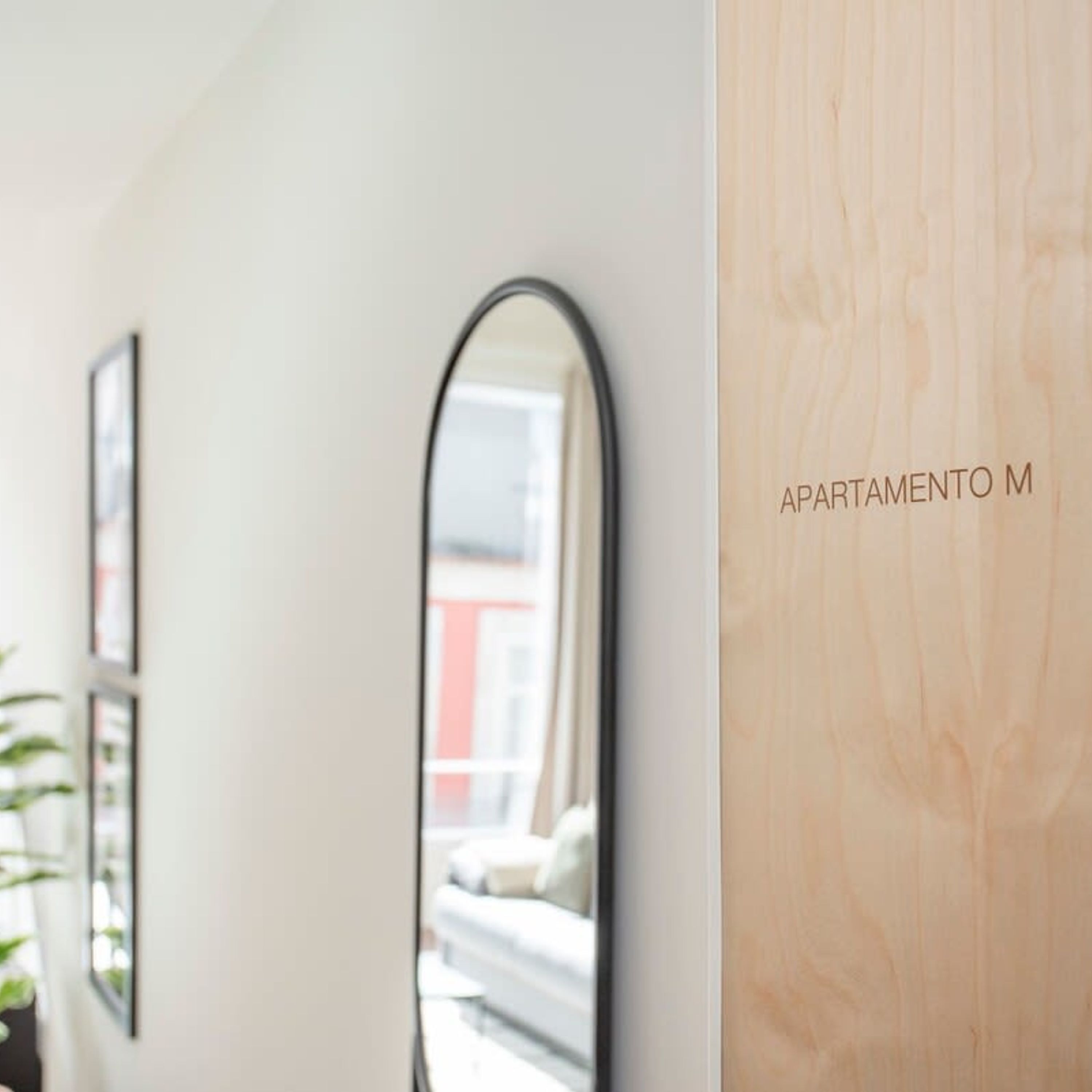 Un couloir avec deux miroirs sur le mur, une porte en bois portant l’inscription « APARTAMENTO M », et, dans le reflet, on aperçoit un lit avec une literie blanche.
