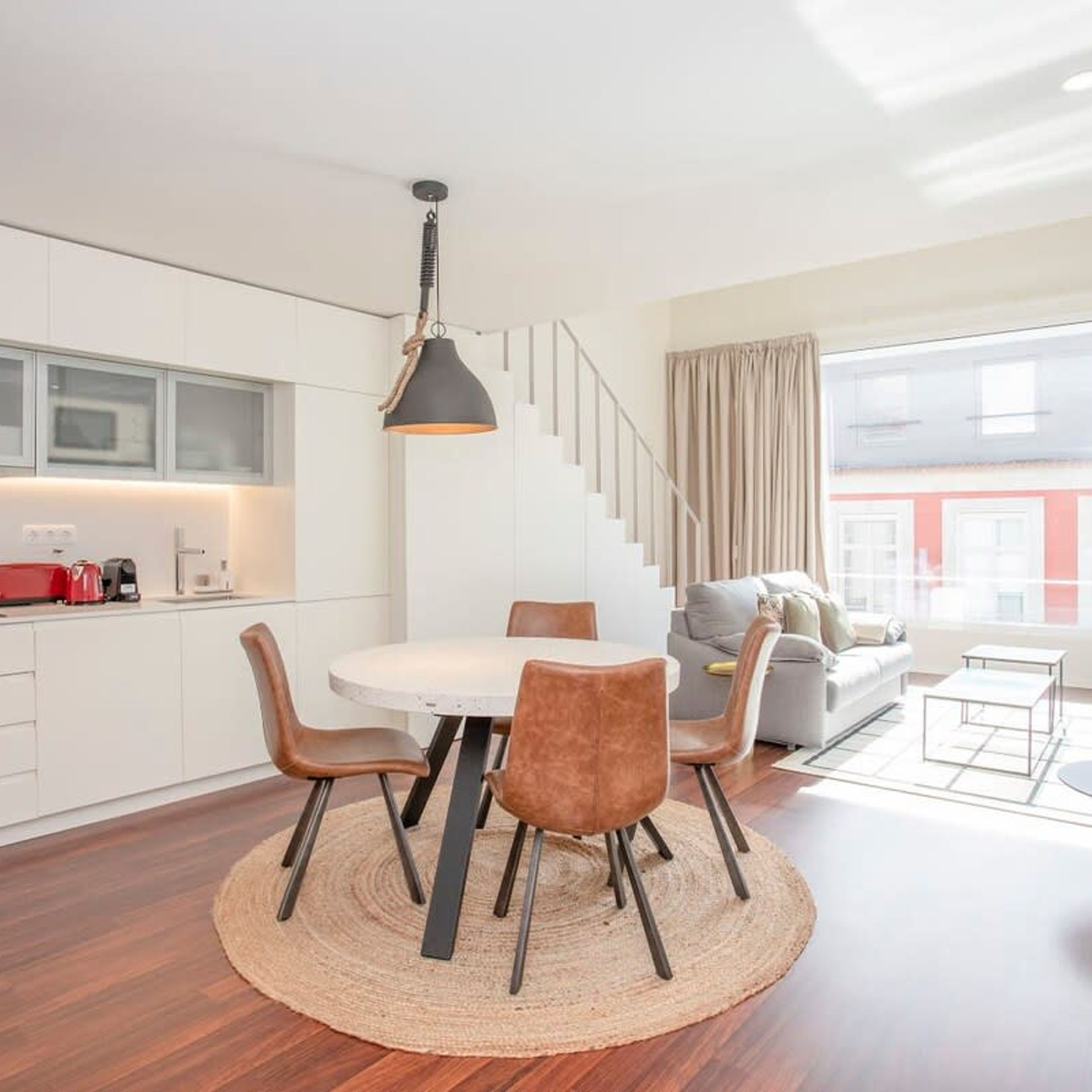 Una cocina moderna con armarios blancos, un comedor con una mesa y sillas redondas, y una escalera que conduce a una sala de estar con sofá y vistas de edificios.