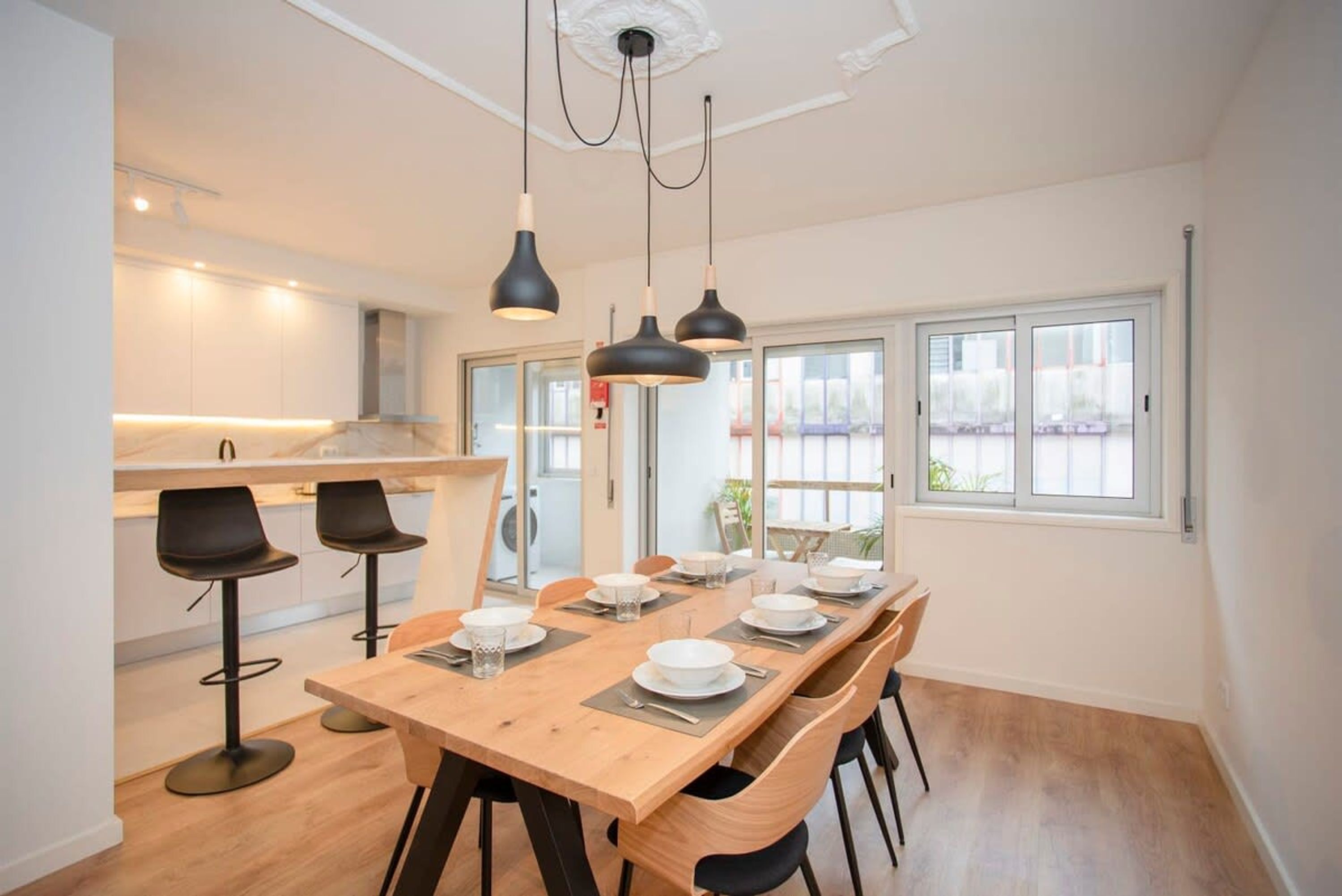 Un comedor moderno con una mesa de madera dispuesta para seis comensales, taburetes negros y lámparas colgantes.