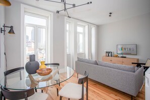 A modern living room with a glass dining table, grey sofa, and a TV on a wooden cabinet. - Liiiving in Porto - Art Design Apartment (Porto)