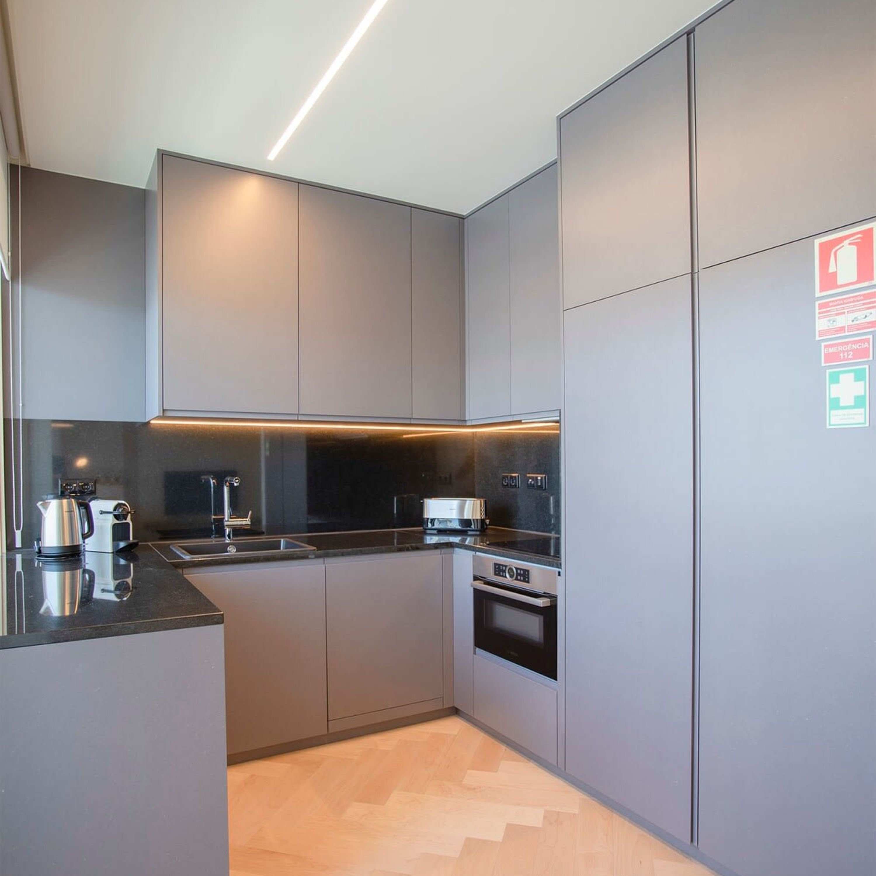A modern kitchen with grey cabinetry, a black countertop, and stainless steel appliances including a kettle, toaster, and oven.