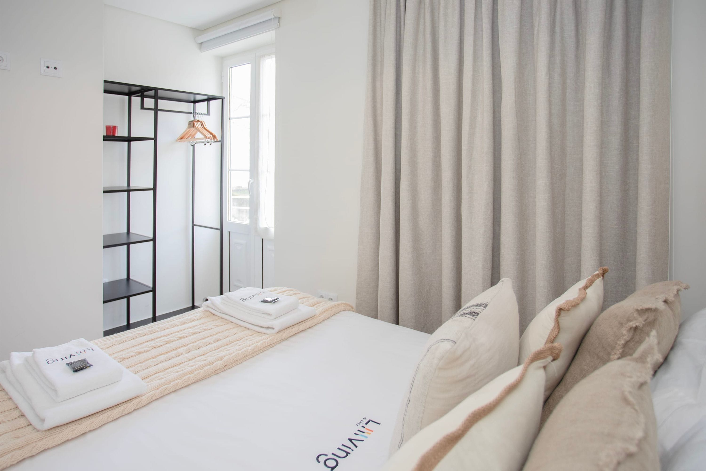 A neatly arranged hotel room with a bed, a hanging coat rack, and a window with curtains.