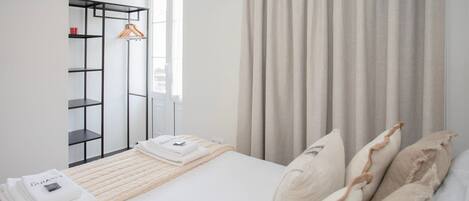 A neatly arranged hotel room with a bed, a hanging coat rack, and a window with curtains.