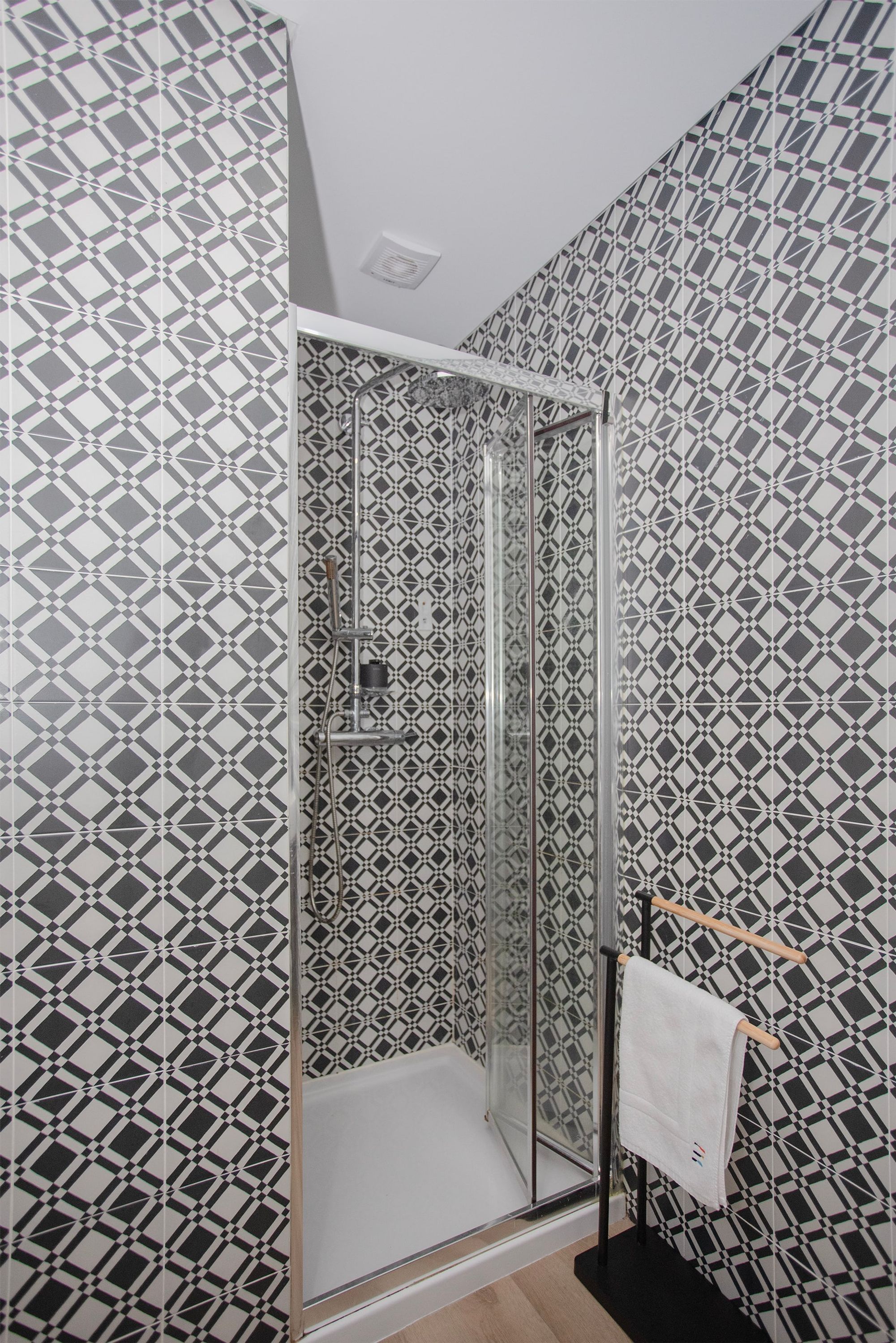 A shower with a geometric patterned wall, a glass door, and a towel rack with a white towel.
