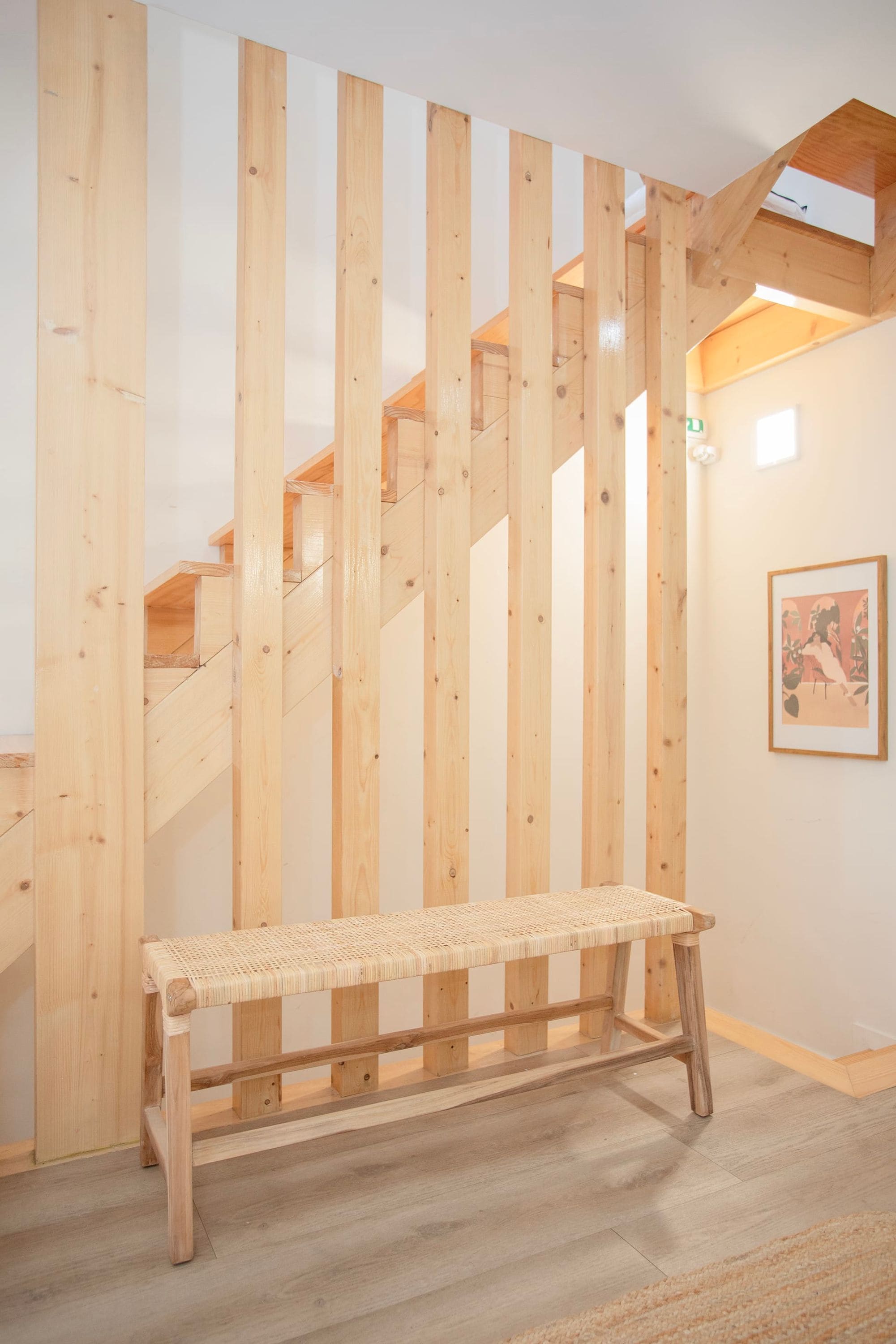 A wooden bench in a room with a wooden staircase and a framed picture on the wall.