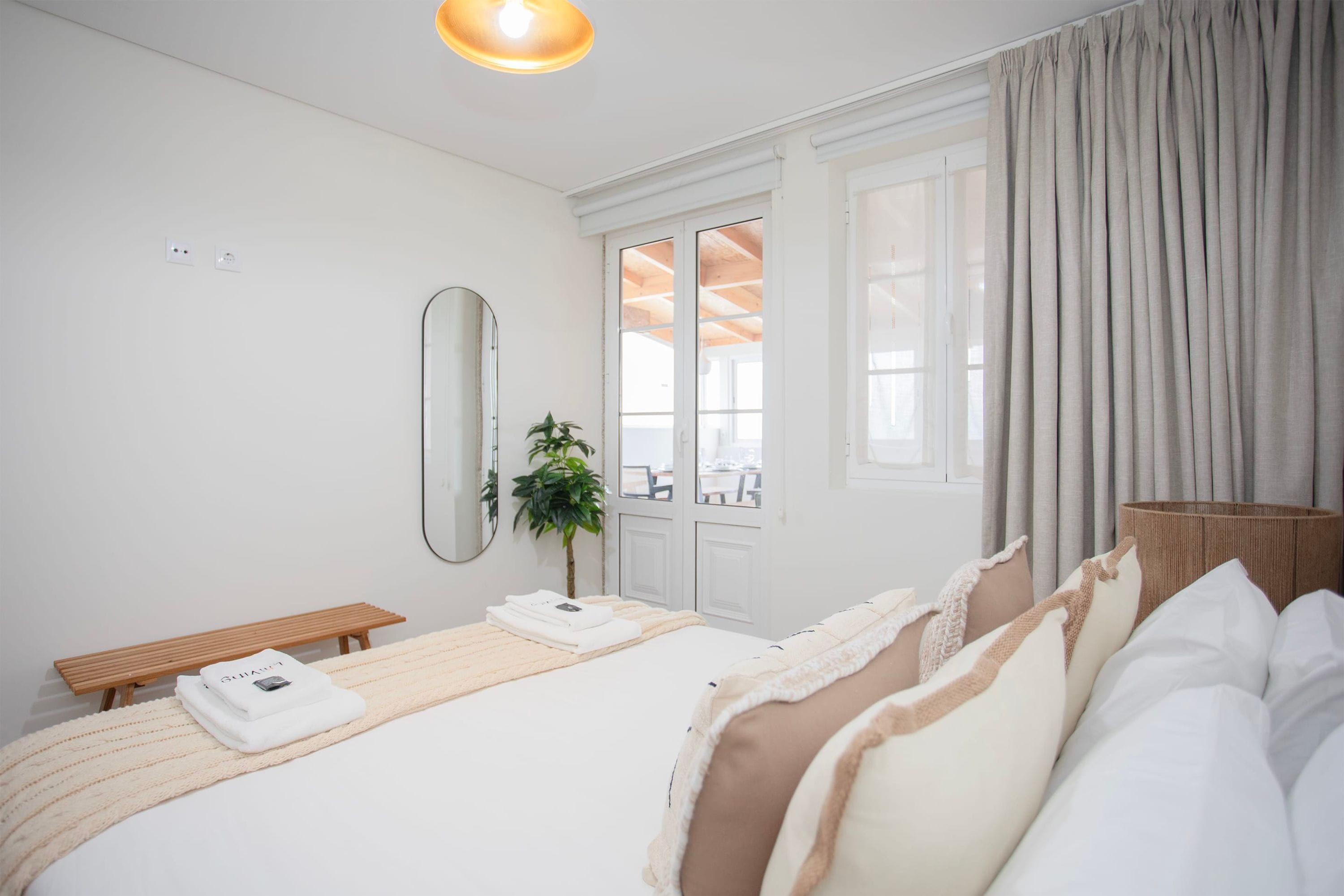 A modern hotel room with a large bed, a bedside table, a mirror, and a view of the outdoors through a window.