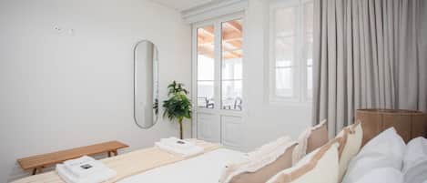 A modern hotel room with a large bed, a bedside table, a mirror, and a view of the outdoors through a window.