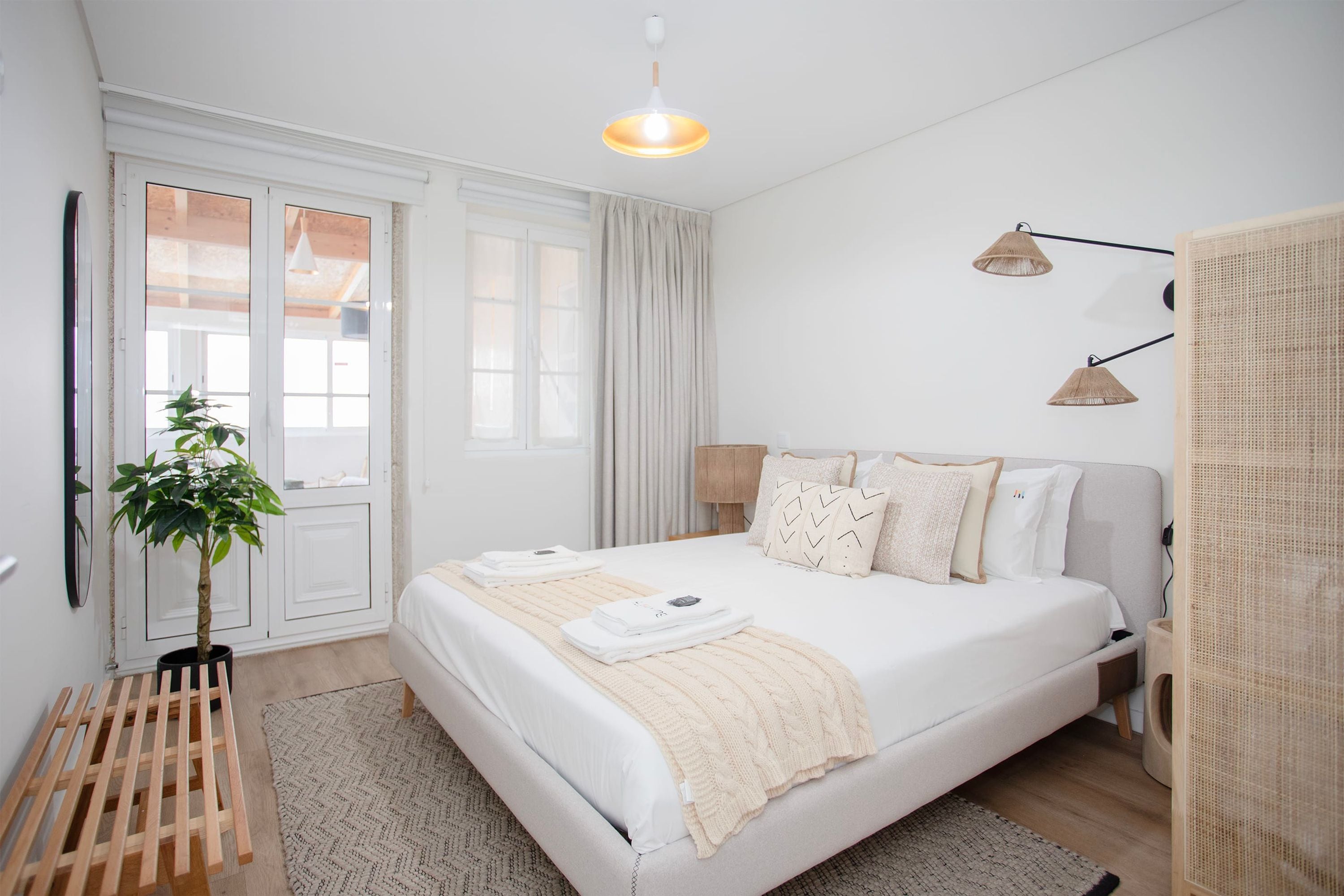 A modern bedroom with a large bed, a bedside table, a lamp, a potted plant, and a window with curtains.